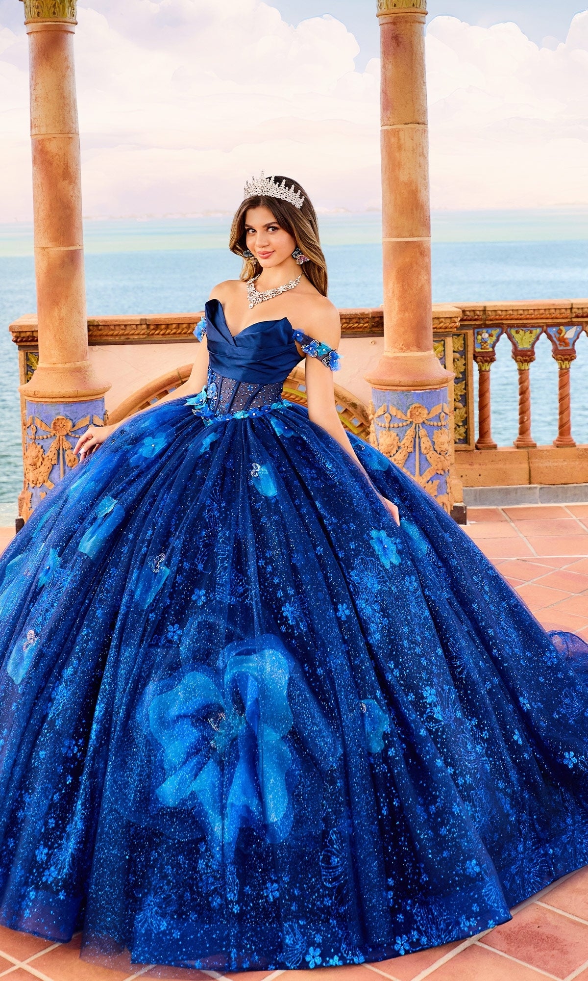 A woman in the Quinceanera Dress PR30159 By Princesa, featuring a blue corset bodice and tiara, stands on a terrace with ornate columns overlooking the sea beneath a partly cloudy sky.